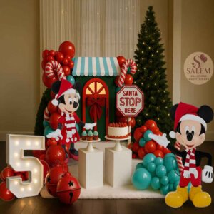 Festive Christmas balloon display featuring a green Santa house with a red bow, surrounded by red and teal balloon garlands, candy cane balloons, a “Santa Stop Here” sign, a lit Christmas tree, a white marquee number 5, large red jingle bells, and two white pedestal stands holding a strawberry cake and cupcakes on a white faux-snow rug. Balloon decor in Oregon.