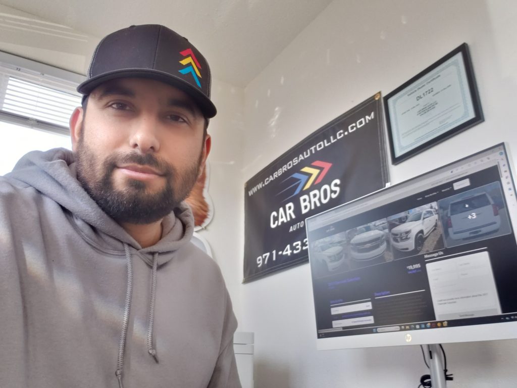Fernando Lopez in his office at Car Bros Auto, LLC in Keizer, Oregon - wearing his car bros logo hat and showing sign on wall as well as computer screen in the background shwoing car for sale.