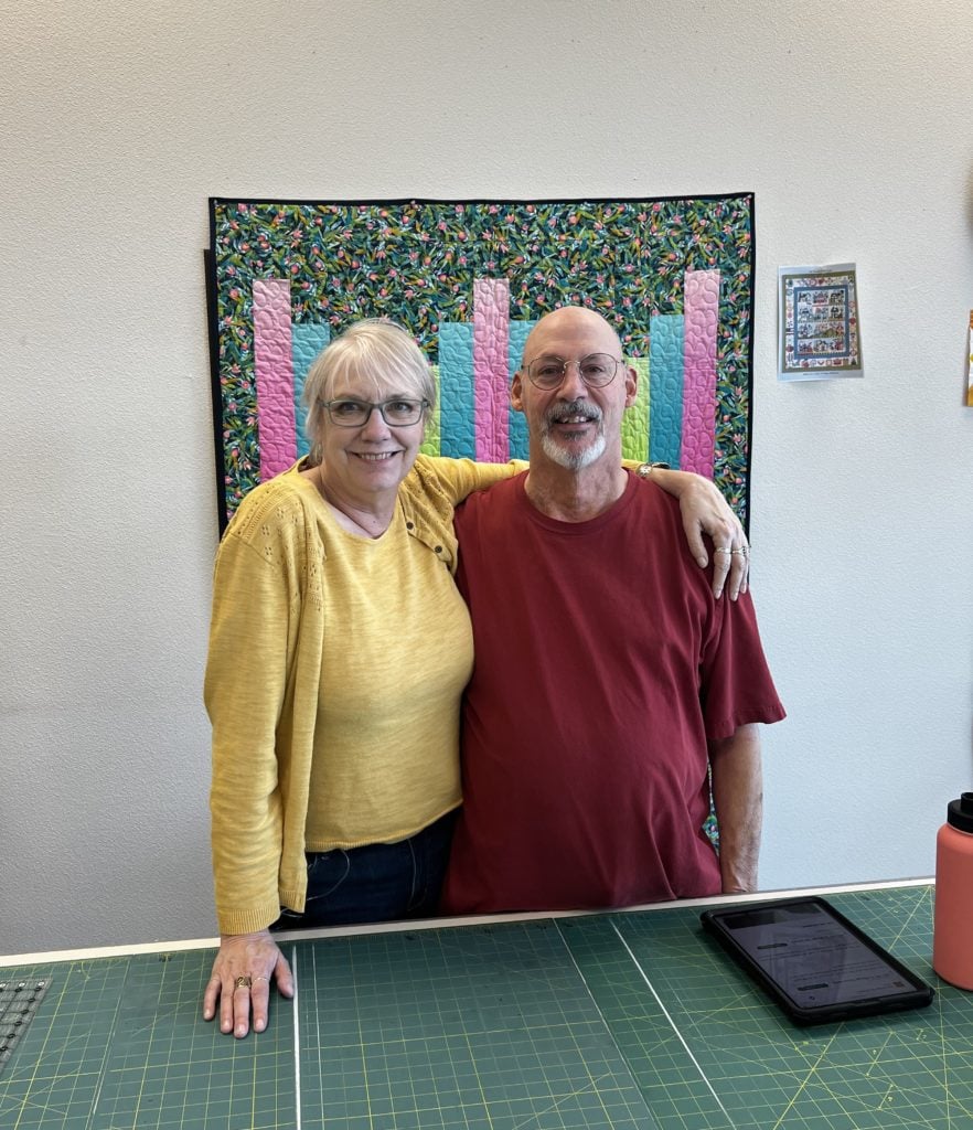 The Cotton Patch Owners wearing yellow and deep red with green gridded cutting mat table and quilt on wall behind.