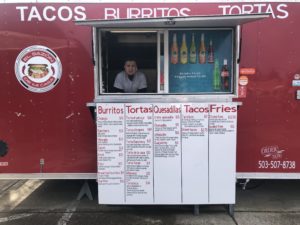 Christian Franco pictured in the window of his food truck facing forward "El Sazon de Mi Casa" in Keizer Orego Deep Red food truck with menu, logo, Tacos, Burritos, Tortas