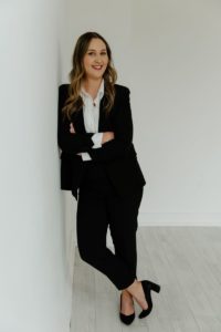 Brooke Hebert, Branch Manager of WaFd Bank, North Salem, Oregon - in a white room with grey wood paneled flooring, wearing a dark suit and white blouse - leaning up against the wall