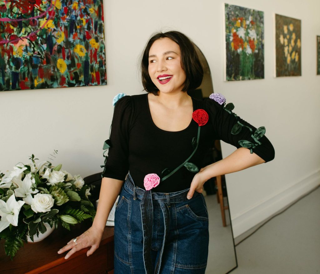 Maria Corpuz of Bel Merch smiles and gazes toward the upper left corner of the frame. A wall lined with artistic pictures stretches out behind her.