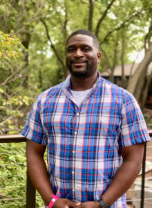 Terril Brown, owner of Uppercuts Barber Shop, stands smiling in the foreground with leafy trees behind him.