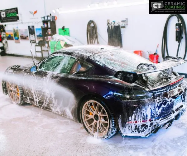 Side-rear view of a dark purple sports car covered in soapy suds during a wash inside a detailing garage.