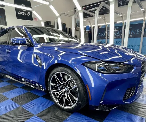 Close-up front angle of the shiny blue BMW sedan showing its custom wheels and reflective finish under bright shop lighting.