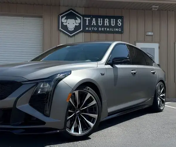 Low-angle, side-profile view of a matte grey Cadillac sedan parked in front of Taurus Auto Detailing.