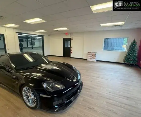 iew of a sleek black Corvette parked on light wood laminate flooring inside a spacious indoor car storage room