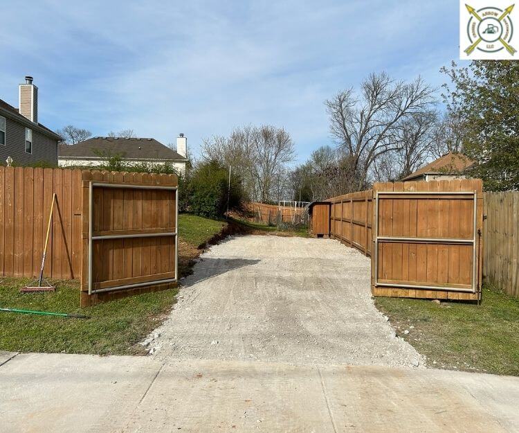 Open wooden fence gates leading into a freshly leveled gravel driveway behind a residential yard.