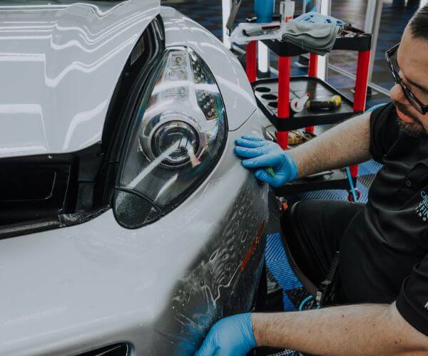 Close-up of a technician applying clear protective film to the front bumper of a white sports car.