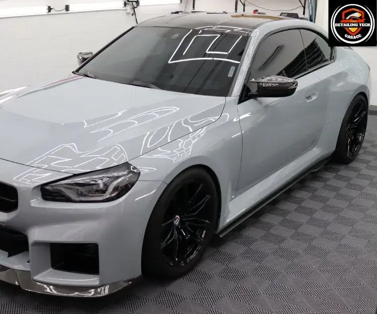 Front three-quarter view of a light gray sports car parked indoors on a checkered floor.